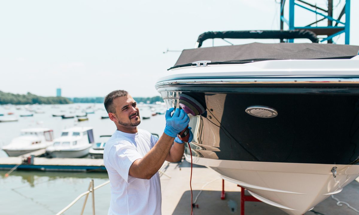 Technician polishing a glossy boat hull with an orbital polisher at a marina